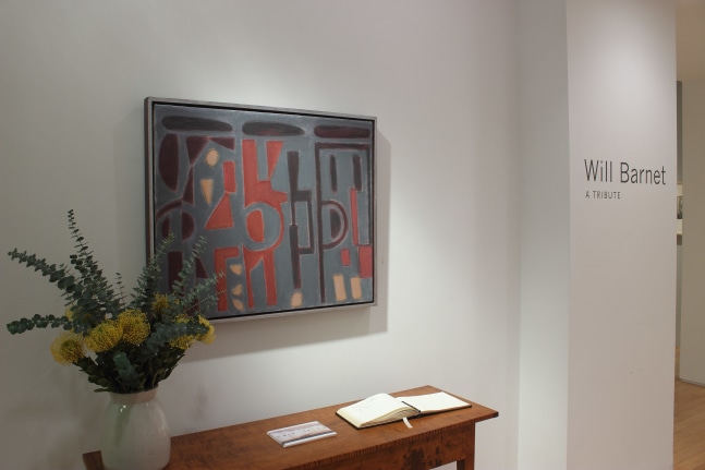 Photograph showing a view of an exhibition with a painting hanging over a wooden table and wall text to the right that reads &quot;Will Barnet: A Tribute&quot;