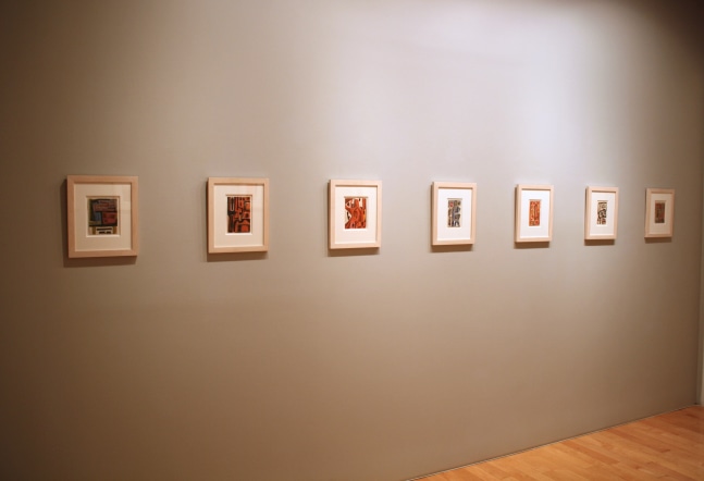 Photograph showing a view of an exhibition space, with seven small framed paintings hanging on a grey wall