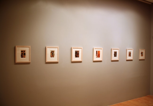 Photograph showing a view of an exhibition space, with seven framed small framed paintings hanging on a grey wall
