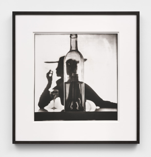 A black and white photographic print by Irving Penn depicting the silhouette of Jean Patchett with a cigarette and wine glass behind a bottle