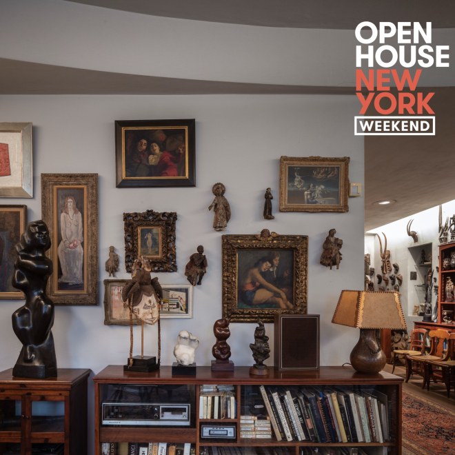 A grey wall of a living room with multiple paintings hung on the walls. There is a short bookshelf in front of the wall with multiple sculptures and a lamp on top. To the right, is part of a hallway lined with chairs, an ornate rug and more sculpture along the walls. Open House New York Weekend is written on the top right corner.