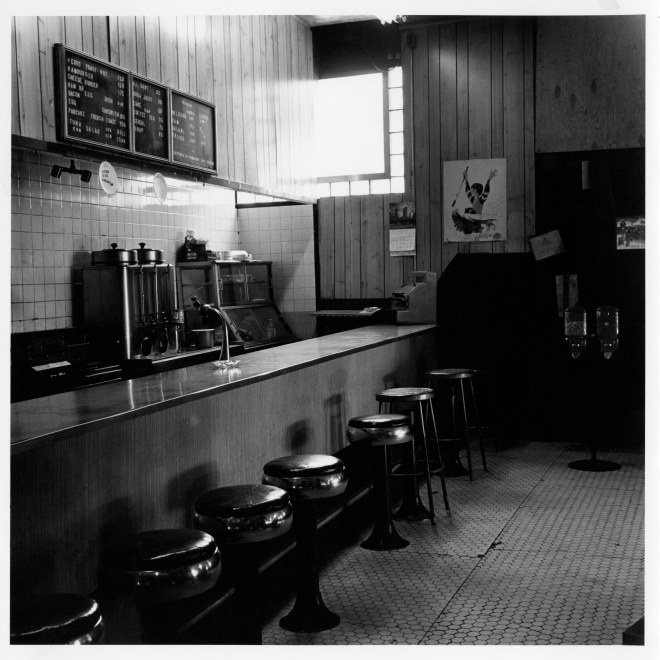 Black and white photograph of an empty bar counter with stools and a tiled floor.