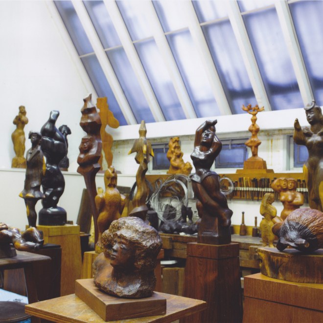 A photo of a high ceilinged, bright, white studio with a large slanted skylight window with a cluster of sculptures in various shades of wood. Almost all of the sculptures depict humanoid faces and abstracted, smooth renditions of the female form. 