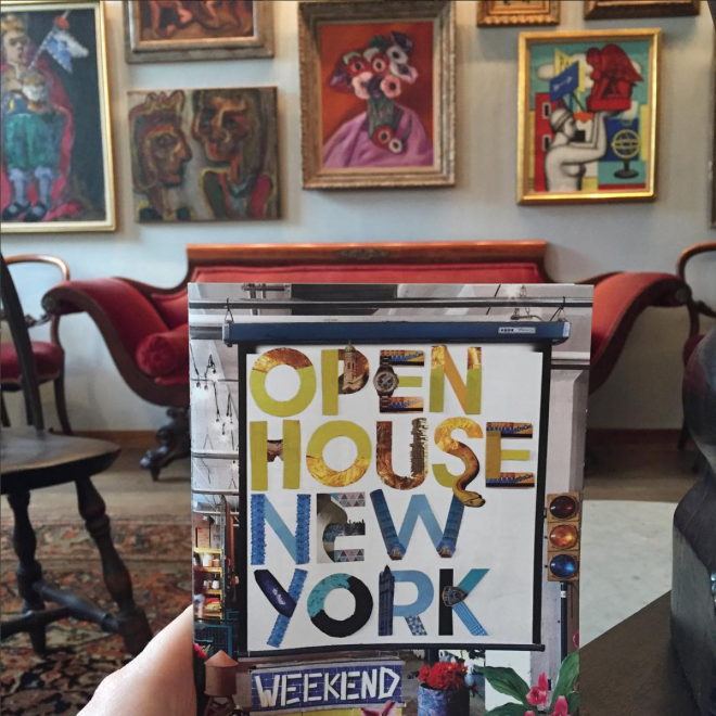 Picture of a pamphlet with the partially visible hand of someone holding it visible. The pamphlet has bright colors, collage styling, and geometric lettering. In the background vibrant pieces of art are framed on the wall and you can seen antique red velvet love seat. 