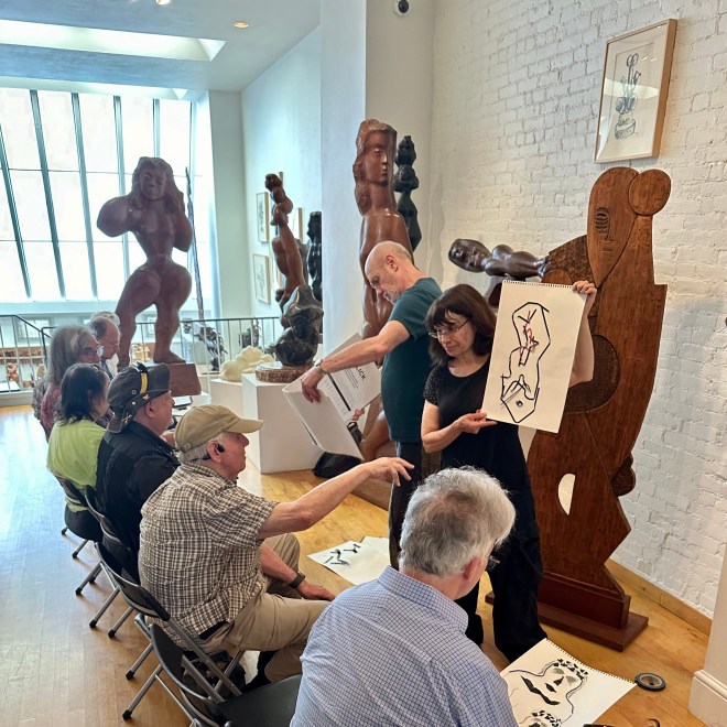 Image of several people sitting down in a long room with sculptures in front of them. Two people also stand in front of the seated group holding up paper with artwork on it.