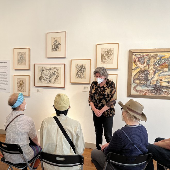A group of people seated in chairs are facing away from the camera towards a woman standing. Behind her on the wall are drawings and watercolors by Chaim Gross.