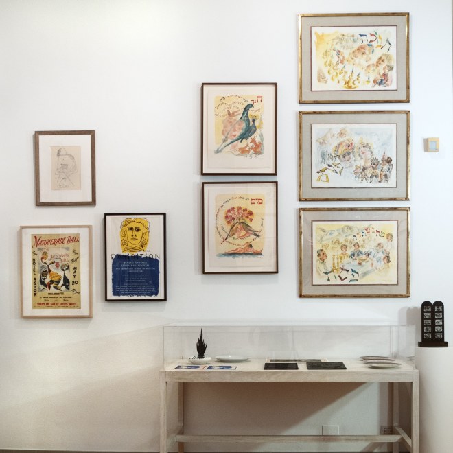 View of exhibition wall with many works in a salon style hang. There is a table in the lower part of the photo with sculpture, ceramics, and two-dimensional works in it.