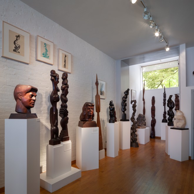 View of a room with a white brick wall on the left and multiple dark brown and black wooden sculptures on pedestals near the walls. There is a window straight ahead near the tall ceiling.