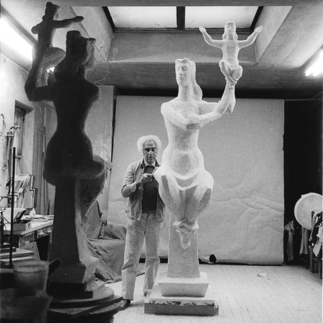 Black and white photograph of a man working on a sculpture in plaster. The sculpture is at the center of the image, with the man to the left. He is working in a room with other sculptures and there is a skylight above him.