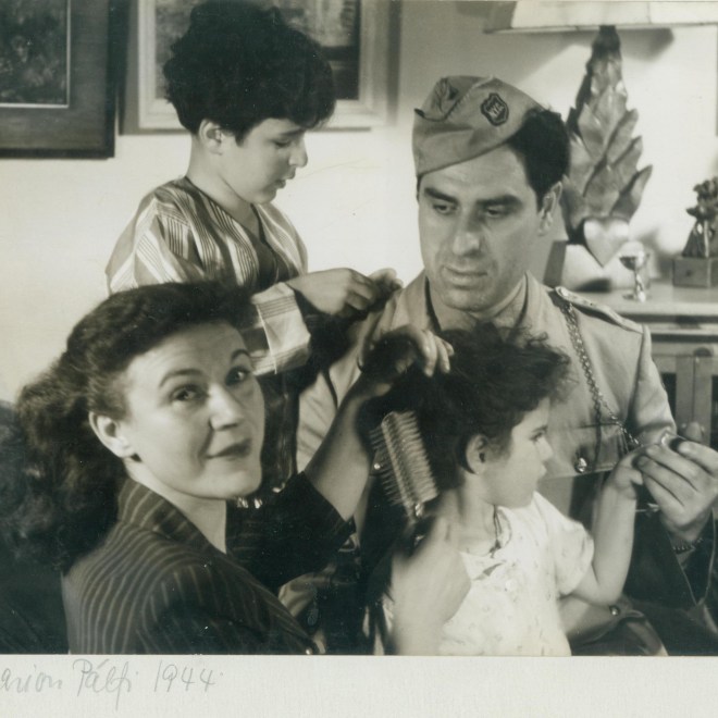 Photo of a family with a husband, wife, boy, and girl. The family is seated close together, with the woman brushing her daughter's hair. The man is in uniform and the children are in pajamas. The woman is the only one looking at the photographer.
