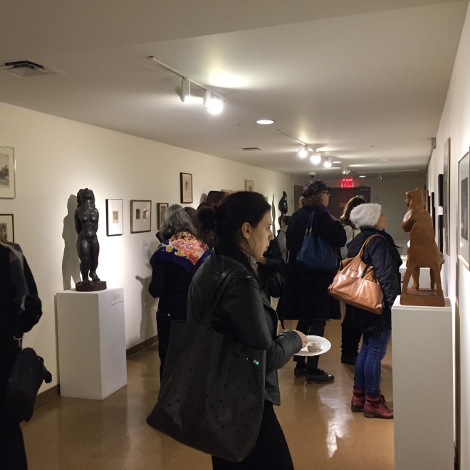 Photo of a gallery with white walls and a light brown floor. The gallery has a low ceiling and adults in dark winter clothes are milling around. On the walls are various pieces of art work in dark brown frames and several large sculptures in shades of brown are visible. To the left of us is a dark colored humanoid sculpture. 