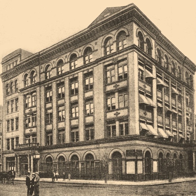 View of three-quarters of a six-story building on a city corner with curved windows and doorways on the top and bottom floors.