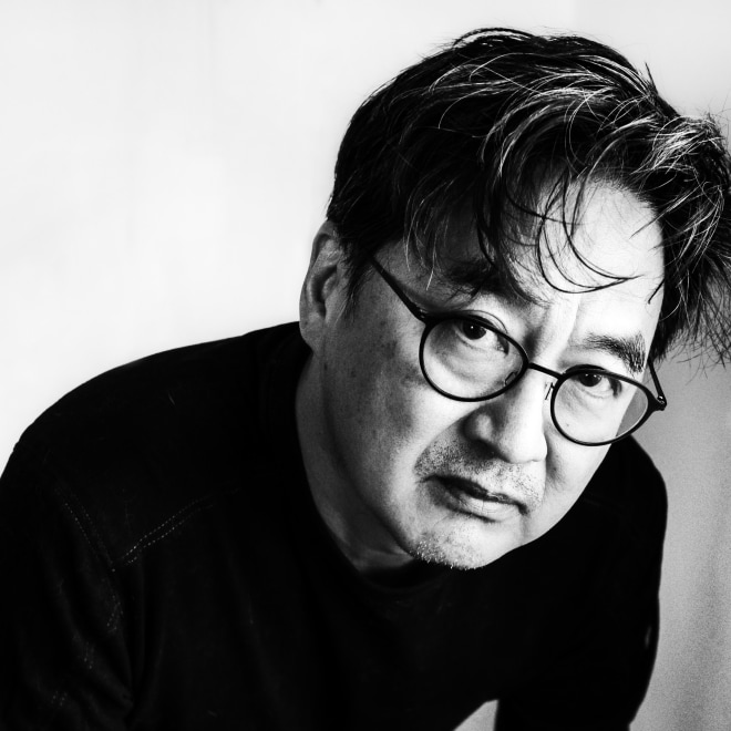 Headshot of a man with glasses and whispy medium length hair. He is leaning towards the camera and is wearing a black shirt.
