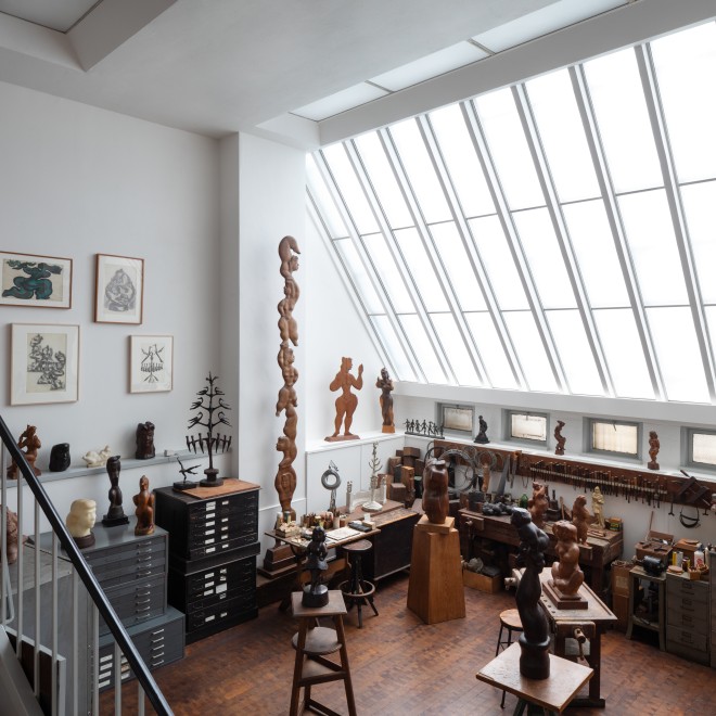 View of Chaim Gross's studio with a large skylight to the right. In the studio are finished sculptures, drawings on the wall, tools, and materials.