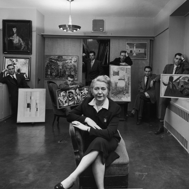 Black and white photo of an older woman seated in the front center, wearing a black suit dress and crossing her legs confidently over her chair. Behind her are six young men, four seated and two standing, wearing dark suits and either holding large pieces of art work or placed near or next to them. 