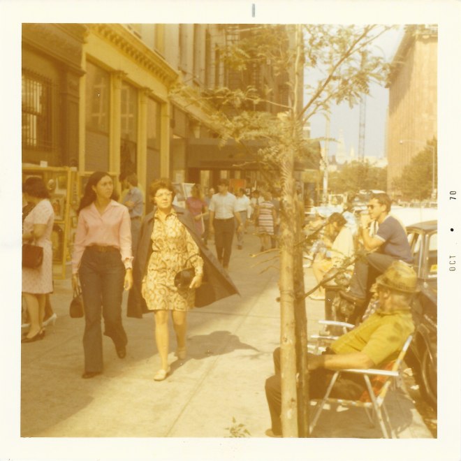 Color snapshot of a city block during the day. There are people selling art and jewelry along the sidewalk. People are walking in the middle of the wide sidewalk, while others are seated in lawn chairs along the street side. 