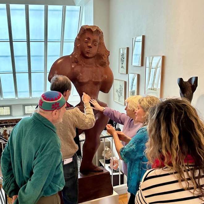 Color image of five people standing near a sculpture of a curvy woman, which stands about 2 feet taller than the people. Two participants are touching the midriff area of the sculpture, while three others wait for their turn to touch. There is a large skylight in the background and drawings on the wall to the right of the sculpture.