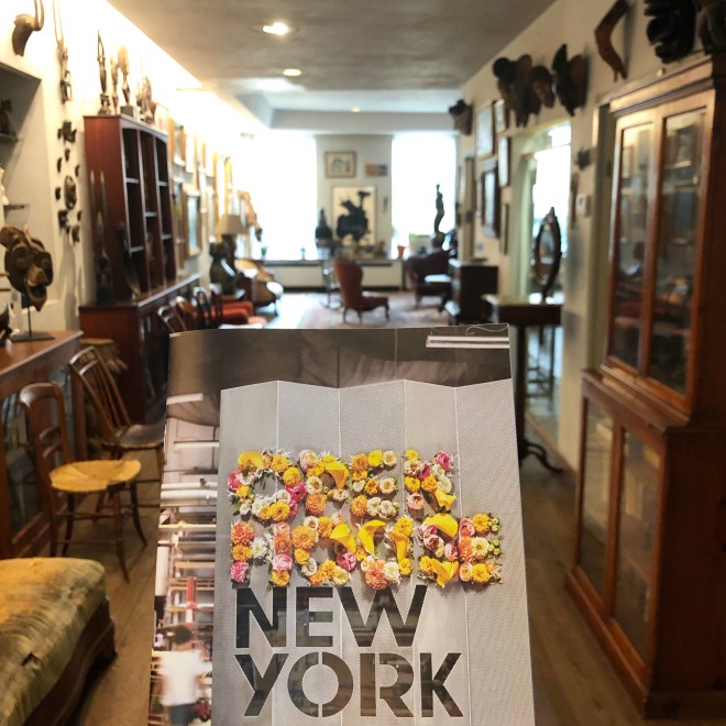 Brochure with bold, black geometric lettering that says New York on the front. In the background is the hallway of a room with dark wood floors and cabinets lining the sides. Sculptures of masks are hanging on the wall to the right and there a chairs scattered about the space. 