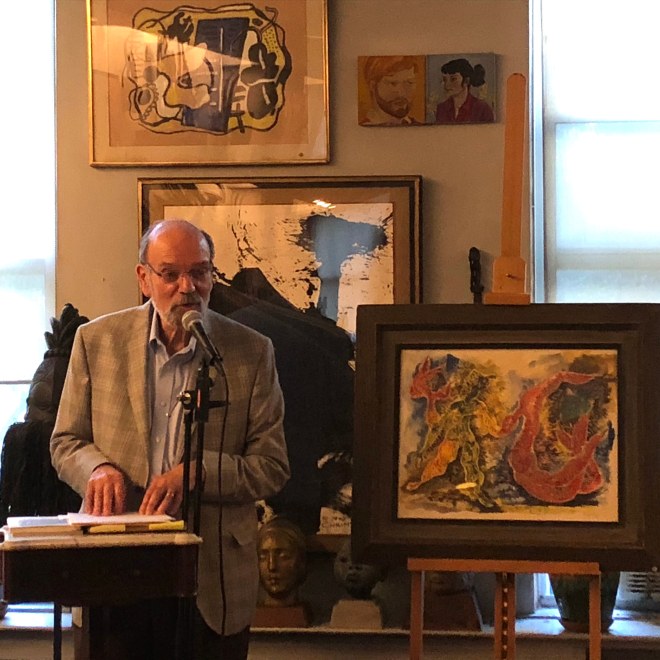 An older man with glasses and a short beard in a grey plaid suit stands to the left of us at a podium speaking into a microphone. To the right of him is a vibrant painting of organic, humanoid forms in a dark brown frame on an easel. 