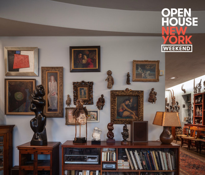 A grey wall of a living room with multiple paintings hung on the walls. There is a short bookshelf in front of the wall with multiple sculptures and a lamp on top. To the right, is part of a hallway lined with chairs, an ornate rug and more sculpture along the walls. Open House New York Weekend is written on the top right corner.