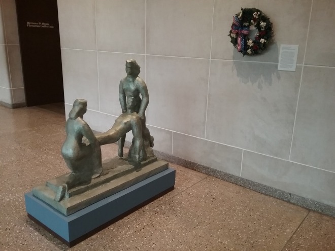 To the left of us is a light grey metal sculpture of two female figures holding the limp body of a male figure between them. Both women have shoulder length hair and the one closest to us is kneeling as she lifts the man. On the wall behind the sculpture is a wreath decorated with white flowers with a bow in the colors of the American flag on it. 