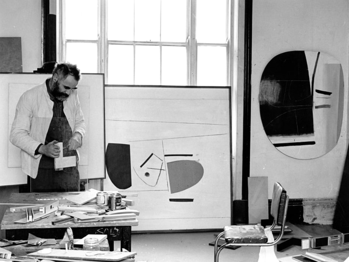 A man with a beard tinkers with wood in a studio surrounded by abstract paintings.