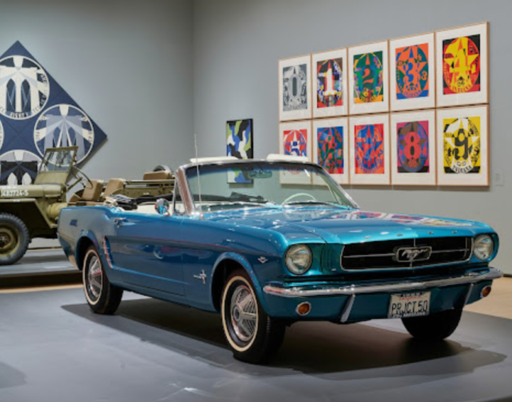 Installation view of Motion. Autos, Art, Architecture, featuring a blue convertible, Indiana's Decade: Autoportrait serigraphs, and his painting The Brooklyn Bridge (only partly visible).