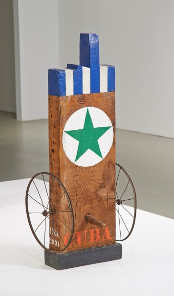 Cuba, a sculpture consisting of a wooden beam with a haunched tenon, standing on a wooden base. The work's title, Cuba, is painted in red letters across the bottom of the plank. Above it is a wooden peg, and to each side is an iron wheel. A green star in a white circle is found in the upper half of the sculpture, and above this the top part of the sculpture, including the tenon, is painted in blue and white vertical stripes.
