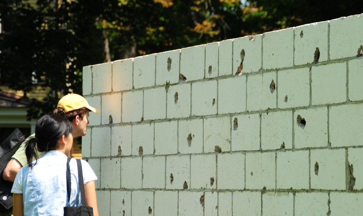 Two people look closely at a fragment of white brick wall with bullet holes