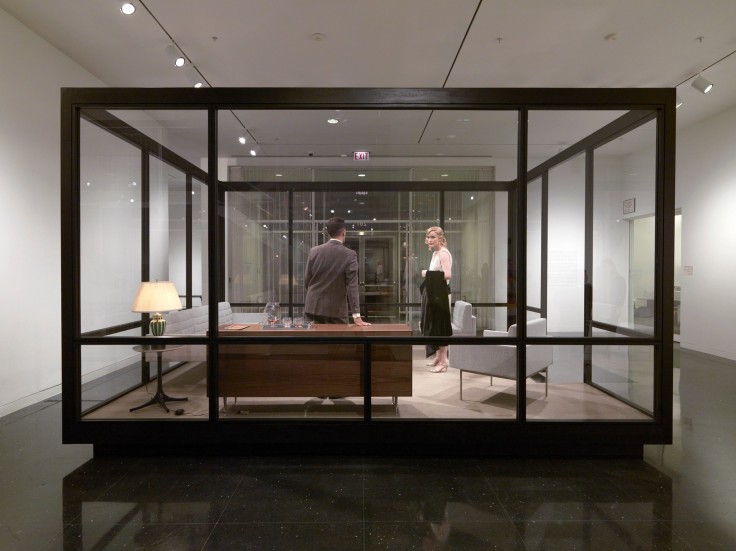 Image of Josiah McElheny's The Club for Modern Fashions,&nbsp;2013 (a giant glass box containing an office space where a man is about to sit on a desk while a woman stares at him)