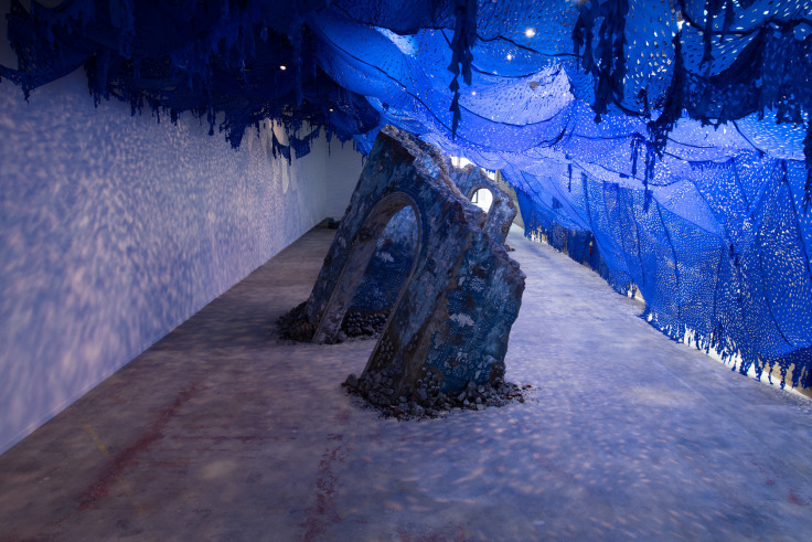 Large sculptural installation of a crumbling blue wall set beneath an indigo-colored textile by Firelei B&aacute;ez.