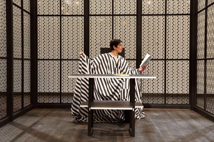 Image of interior view of Josiah McElheny's The Ornament Museum, 2016 (woman wearing a black and white striped tunic seated while holding and reading a document)