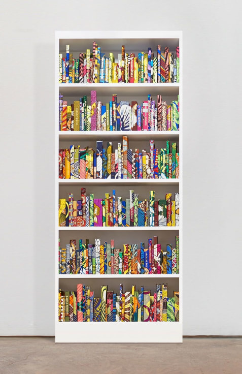 White wooden bookshelf containing approximately 225 hardback books bound in Dutch wax print cotton textile with names embossed in gold on their spines