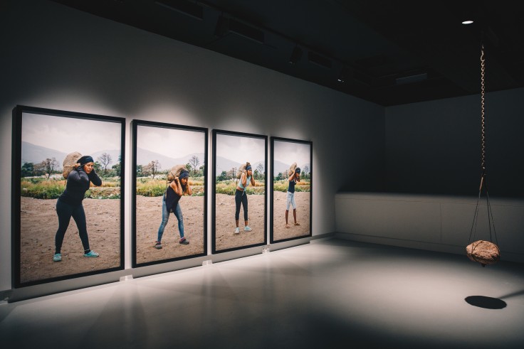 Four framed photographs of female labor carrying stones on their shoulders, facing at a strapped stone installation