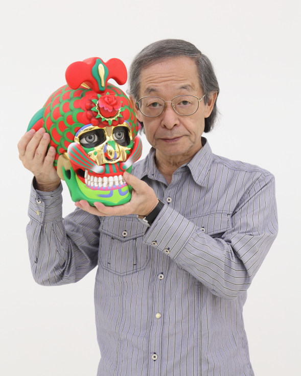 Portrait of artist Keiichi Tanaami holding one of his brightly colored sculptures that looks like a skull