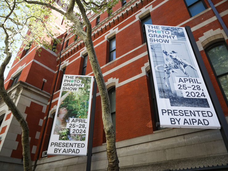 Two banners with photographs and text advertising The Photography Show 2024 are shown outside the Park Avenue Armory with trees in front of the building as well.
