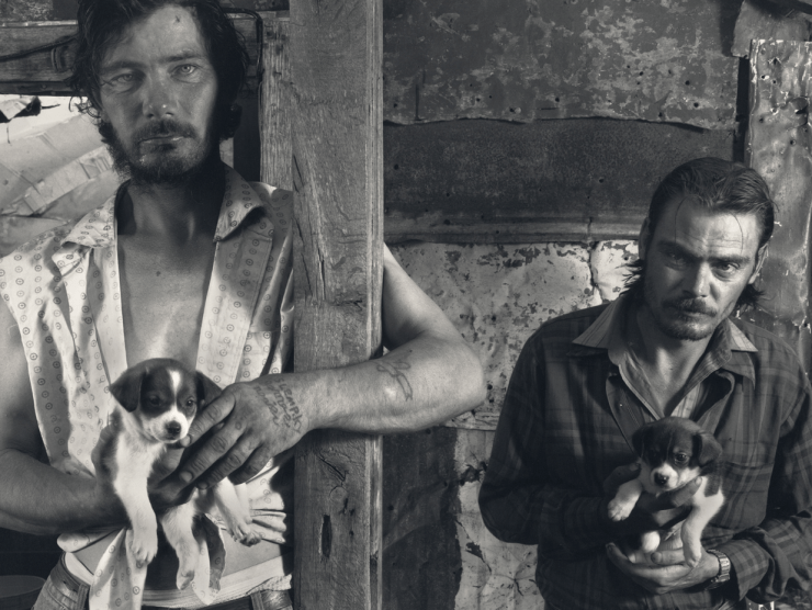 Two rugged-looking white men look at the camera while each holding an adorable puppy in this black and white photograph.