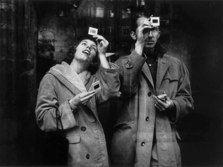 A man and a woman photographed in black and white hold up slides and look through them to see the image
