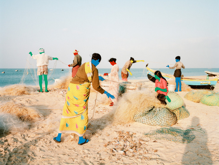 A group of people fish by the sea shore, holding nets, but their skin has been changed to bright primary colors such as blue, green, and red.