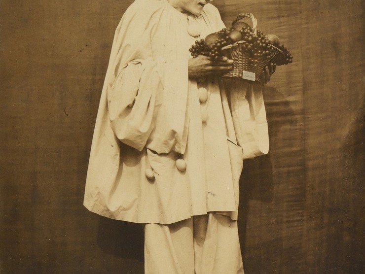 Pierrot the clown holds a basket of fruit while looking down at it in this sepia photograph.