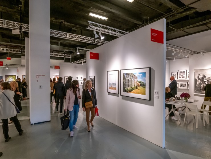 Two women walk through an aisle of The Photography Show by a bright and colorful photograph.