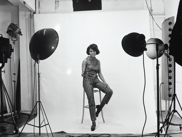 A young Black woman sits on a stool in a photo studio, while smiling and holding a light meter in her hand.