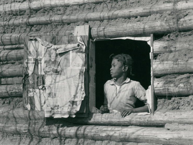 A Black child looks out a window of a log house which has newspapers on the window shutter