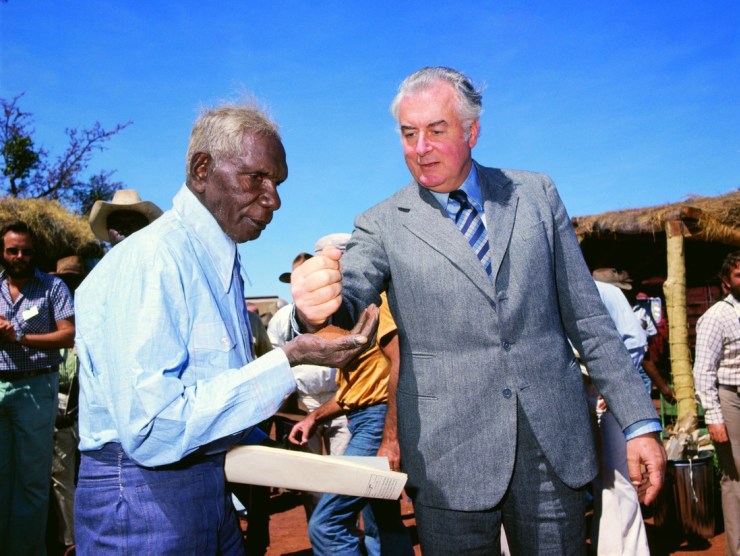 An older white man pours earth into the palm of an older black man.