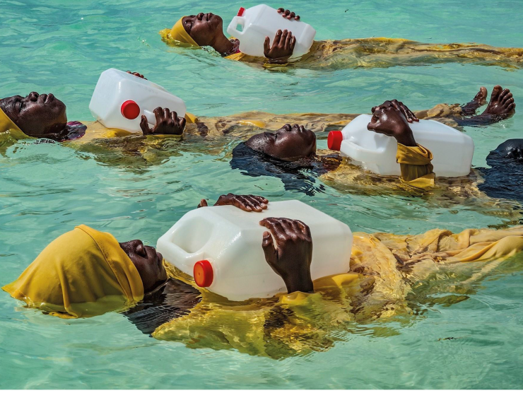 Four Black women wearing yellow clothes and headscarves float in bright turquoise water while holding plastic jugs.