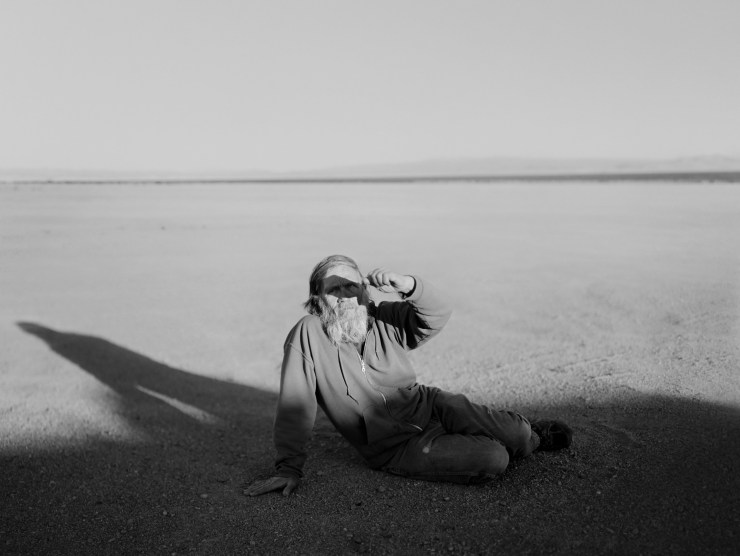 A man is photographed in black and white in a desolate desert-like landscape, as he sits on the ground and shields his eyes from the sun.