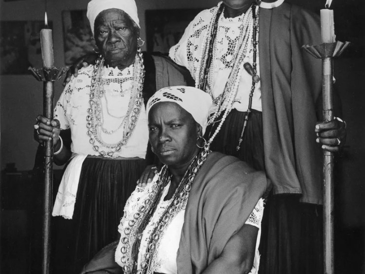 Three Black elders wear ceremonial dress and two hold candles