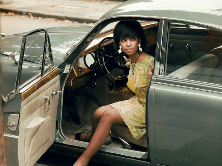 A young Black woman sits in the driver's seat of a shiny olive green car and looks out in the distance.