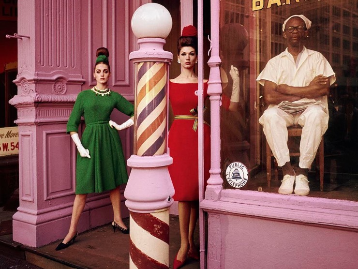 Two white female models stand in brightly colored clothing in front of a pink barbershop while the Black male barber sits in the storefront on a chair and all three people look at the camera.