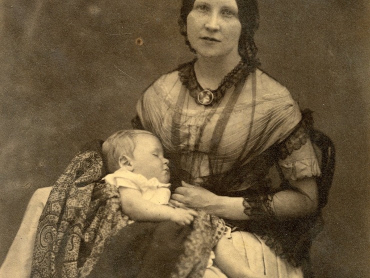 A black and white sepia photo of a seated woman dressed in formal clothes and holding a baby.